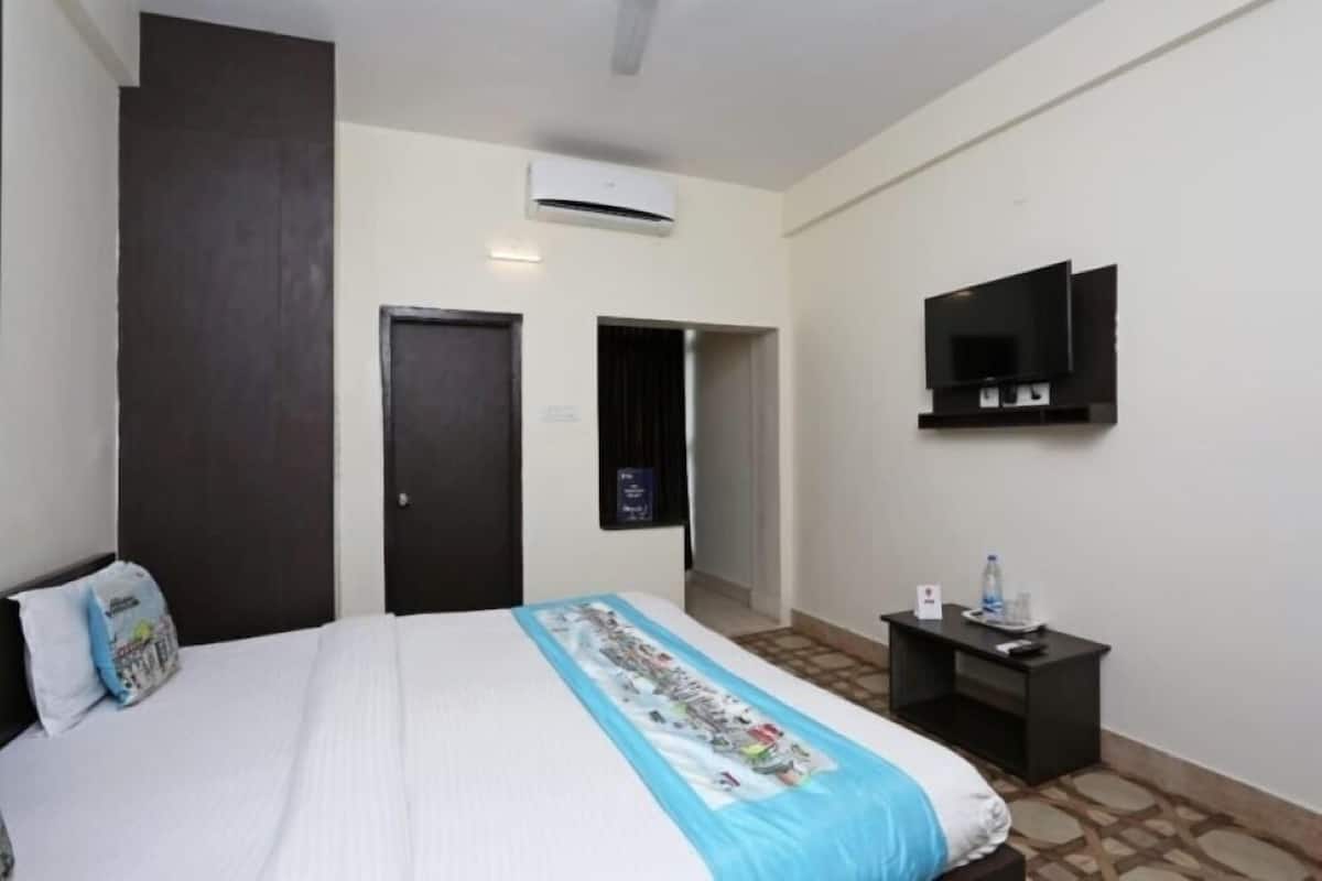 A hotel room with a bed, a TV mounted on the wall, a small table with a bottle and glasses, and an air conditioning unit.
