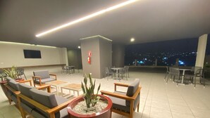 Un salón en la azotea con arreglos de asientos, una planta en maceta y vistas nocturnas de la ciudad.