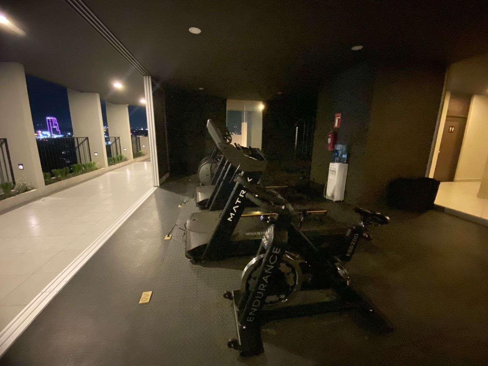 Een sportschool met loopbanden en hometrainers, een waterfontein en uitzicht op de stad 's nachts.