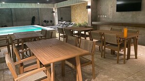 Un área junto a la piscina con mesas y sillas de madera, un muro de piedra y un televisor de pantalla plana montado en la pared.