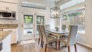 Uma cozinha moderna com micro-ondas embutido, uma sala de jantar com mesa de madeira e cadeiras estofadas, e vista para um quintal através de portas de vidro.