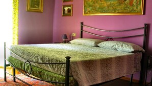 Un letto con struttura in metallo, una coperta a motivi decorativi e cuscini. È presente un comodino con una lampada e un quadro appeso al muro.