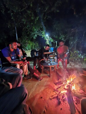 Un gruppo di persone sedute attorno a un fuoco da campo di notte, con un tavolo e delle sedie nelle vicinanze.
