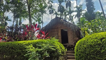 A thatched-roof structure with a staircase leading to an entrance, surrounded by lush greenery and palm trees.
