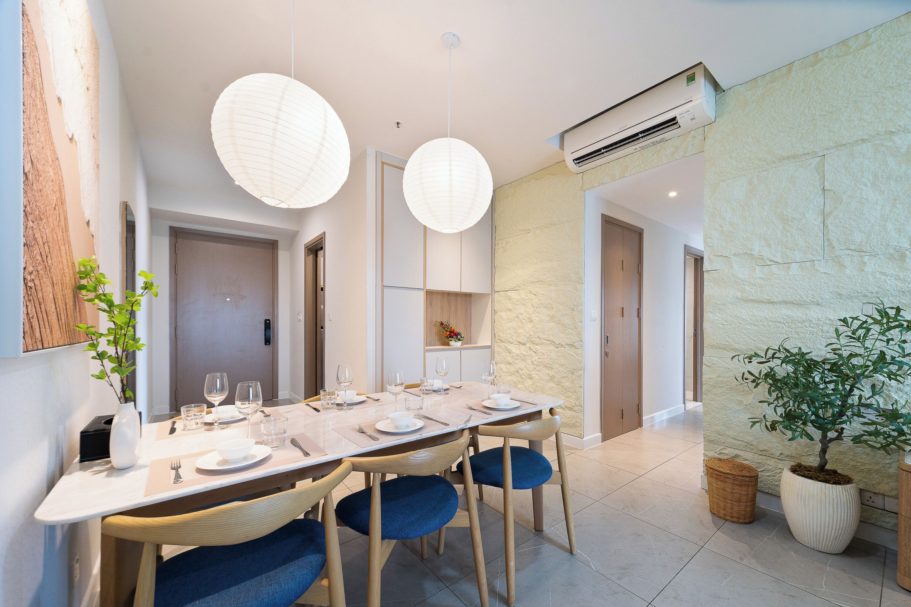 A dining area with a long table set for six, surrounded by chairs with blue cushions, a vase with a plant, and two hanging paper lanterns.