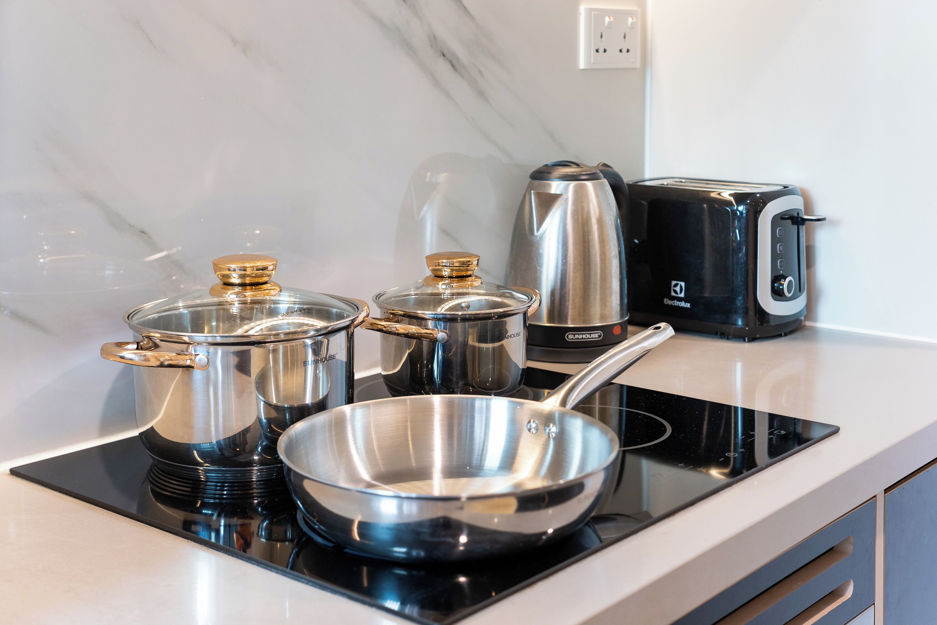 A kitchen with a black induction cooktop, stainless steel pots, a frying pan, a kettle, and a toaster.
