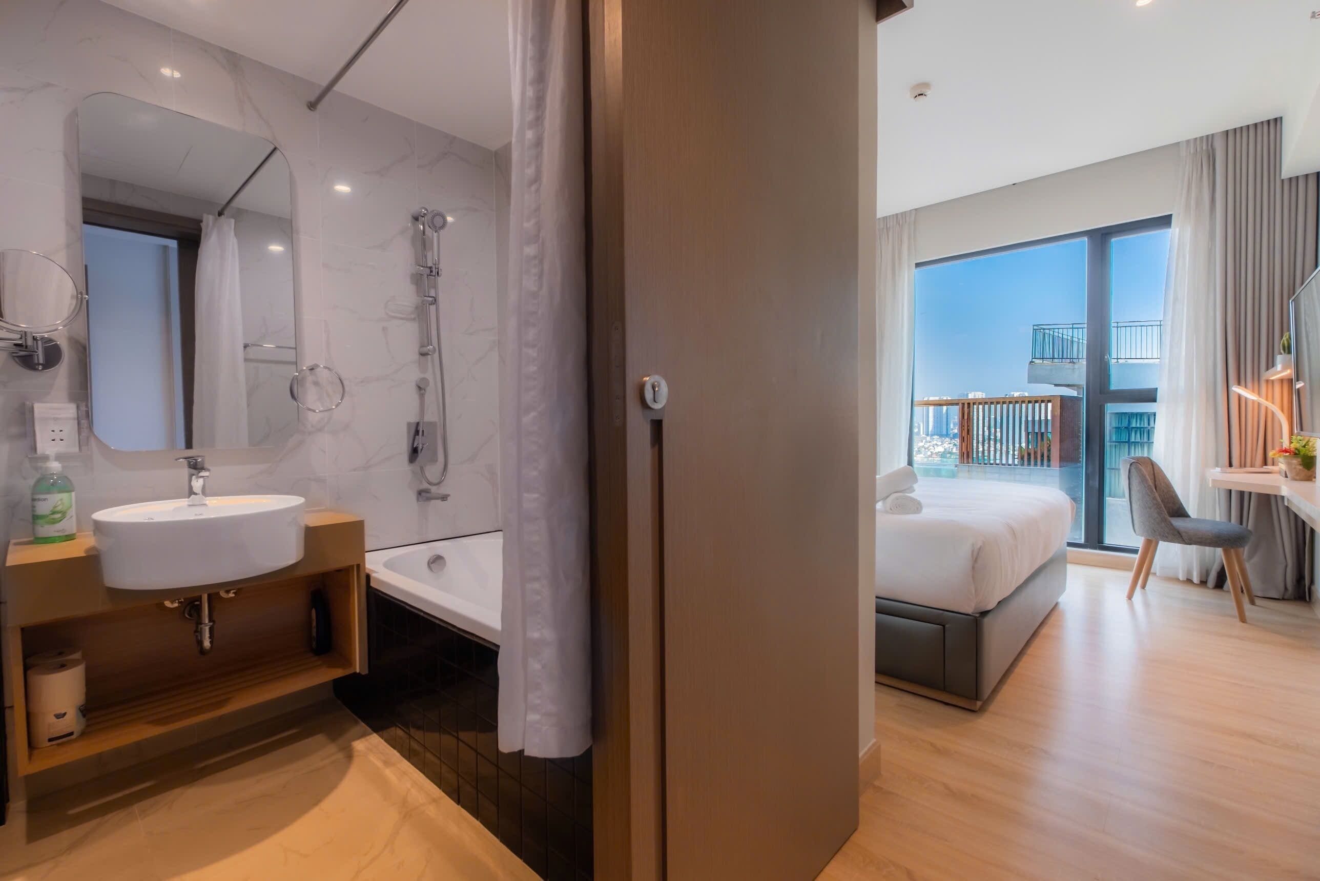 A modern hotel room with a bathroom featuring a sink, mirror, and shower. The room has a balcony with a view of the city.