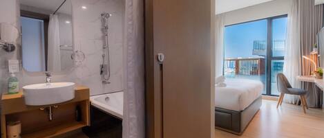 A modern hotel room with a bathroom featuring a sink, mirror, and shower. The room has a balcony with a view of the city.