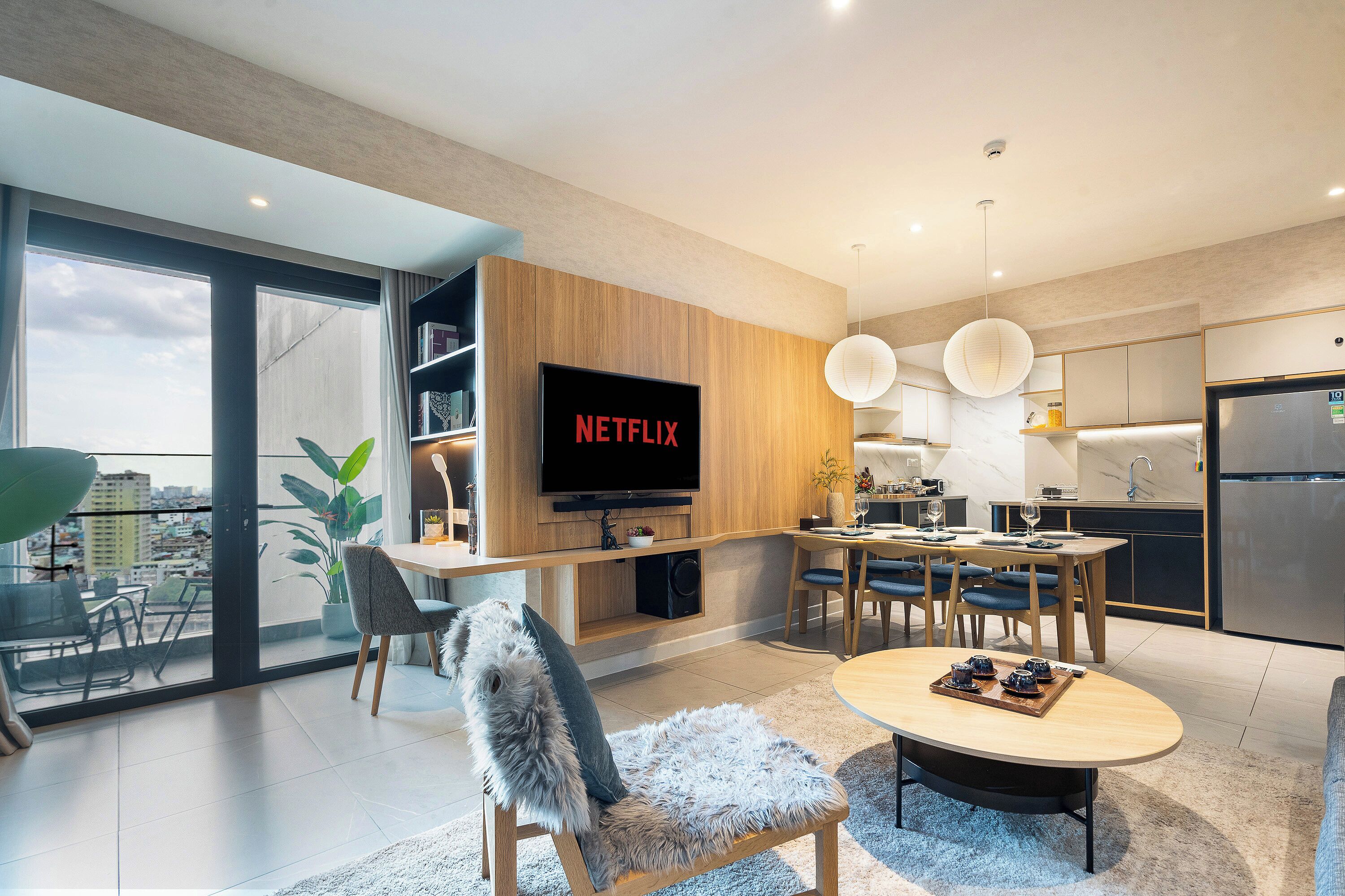 A modern living room with a large flat-screen TV, a cozy seating area, a dining space, and a kitchen with a stainless steel refrigerator.