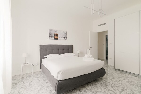 A modern hotel room with a large bed, a small bedside table, a lamp, a towel rack, and a framed picture on the wall.