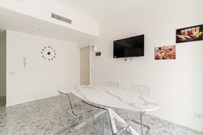 Une pièce moderne avec une table en marbre, des chaises transparentes, un téléviseur fixé au mur et une horloge murale.