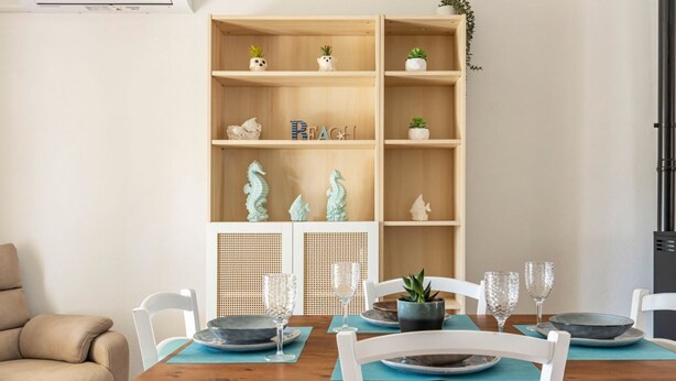 A dining area with a wooden table, white chairs, and a teal-colored table runner. There are decorative items on shelves behind the table, including a sign that reads 'PEACH.'