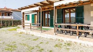 A building with a covered outdoor seating area, a door with green shutters, and a gravel ground.