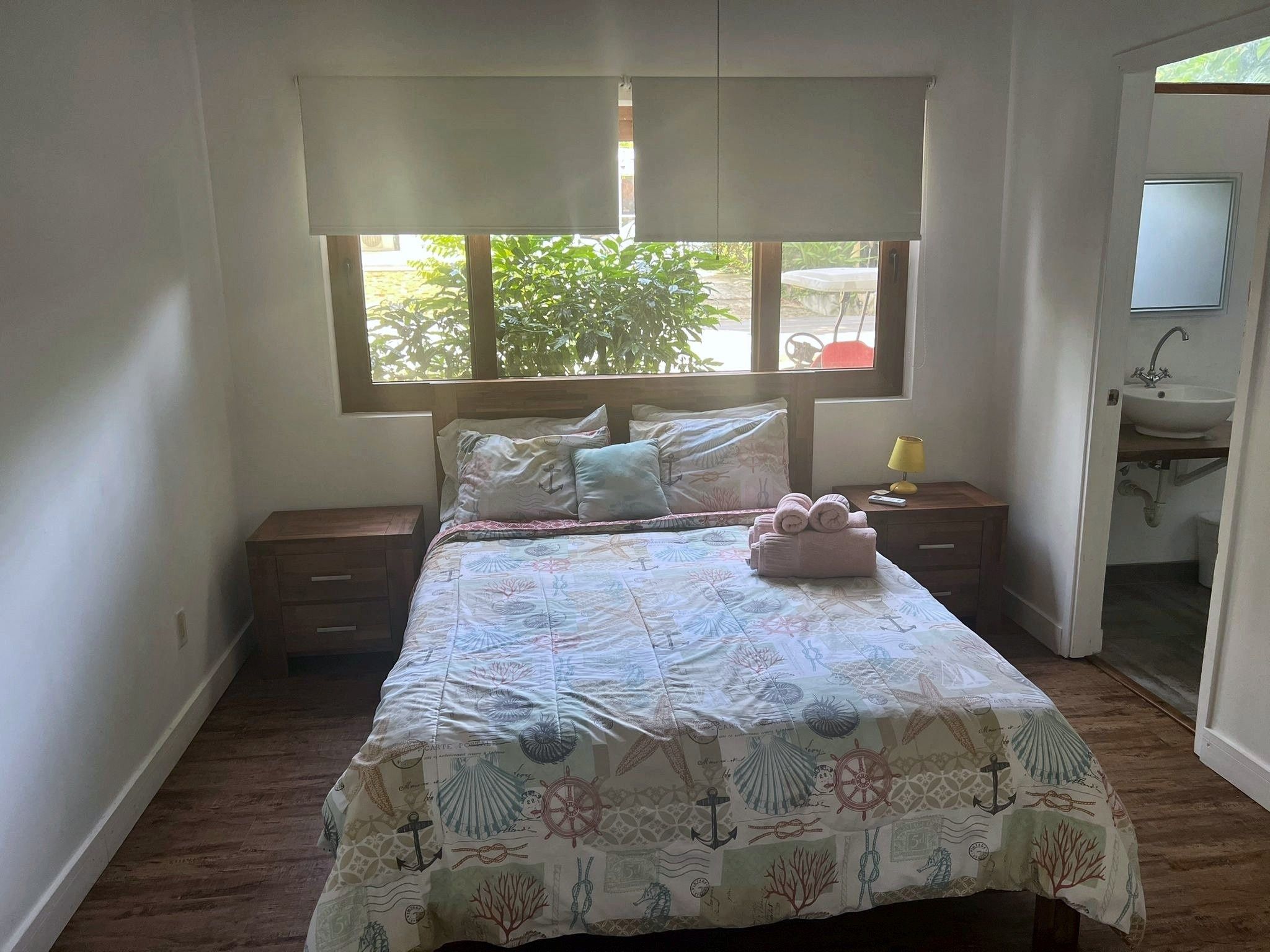 A bedroom with a bed, a nightstand, a window with blinds, and a view of the outdoors.