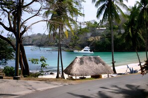 A beach with a thatched-roof hut, a white boat on the water, and palm trees. - Gemini Villas Gemini I (Saboga)
