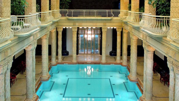 Una gran piscina interior con una isla central, rodeada de columnas ornamentales y un balcón con barandilla.