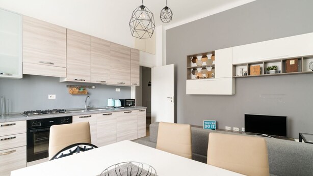 Een moderne keuken met lichte houten kastjes, een eettafel met stoelen, en een grijze bank in de woonkamer.