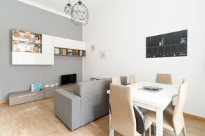A modern living room with a grey sofa, a white dining table with chairs, a TV, and a geometric pendant light.
