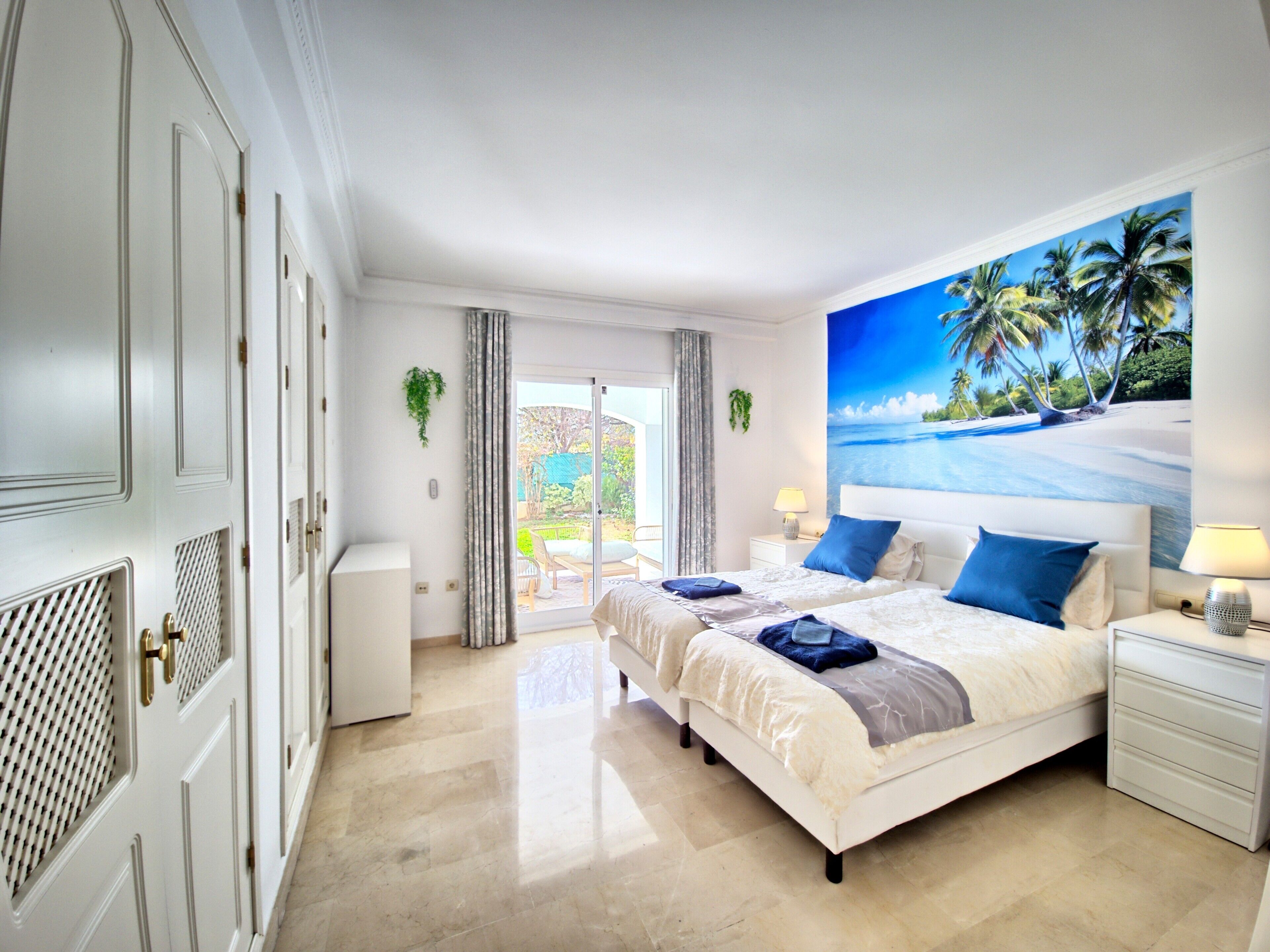 A hotel room with two beds, a large beach mural, a sliding glass door to the balcony, and a white wardrobe.