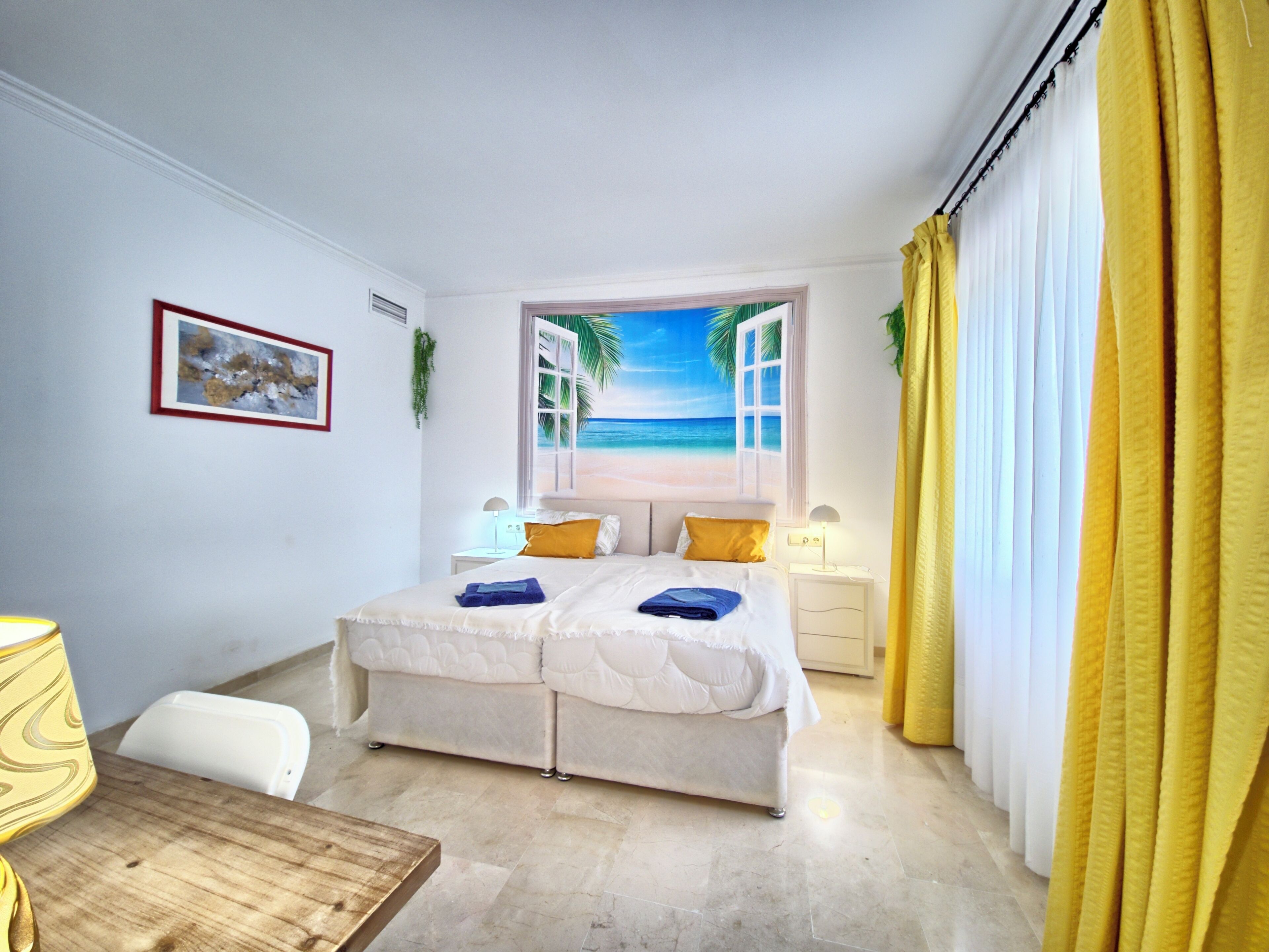 A bedroom with a bed, bedside tables, a nightstand, a window with a beach view, and curtains.