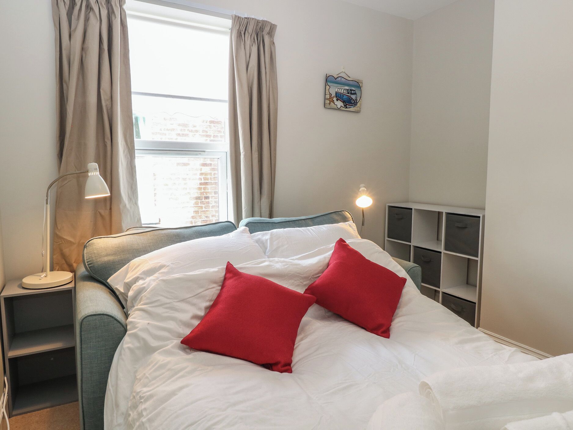 A bedroom with a bed, two red pillows, a bedside lamp, a window with curtains, and a wall-mounted shelf.