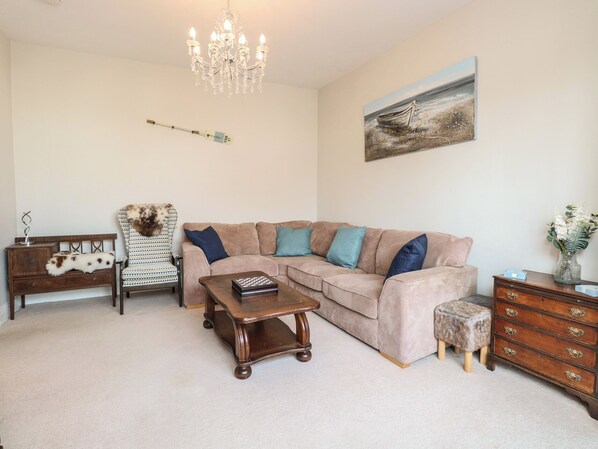 A living room with a beige sectional sofa, a wooden coffee table, a patterned armchair, a small wooden side table, and a chandelier.