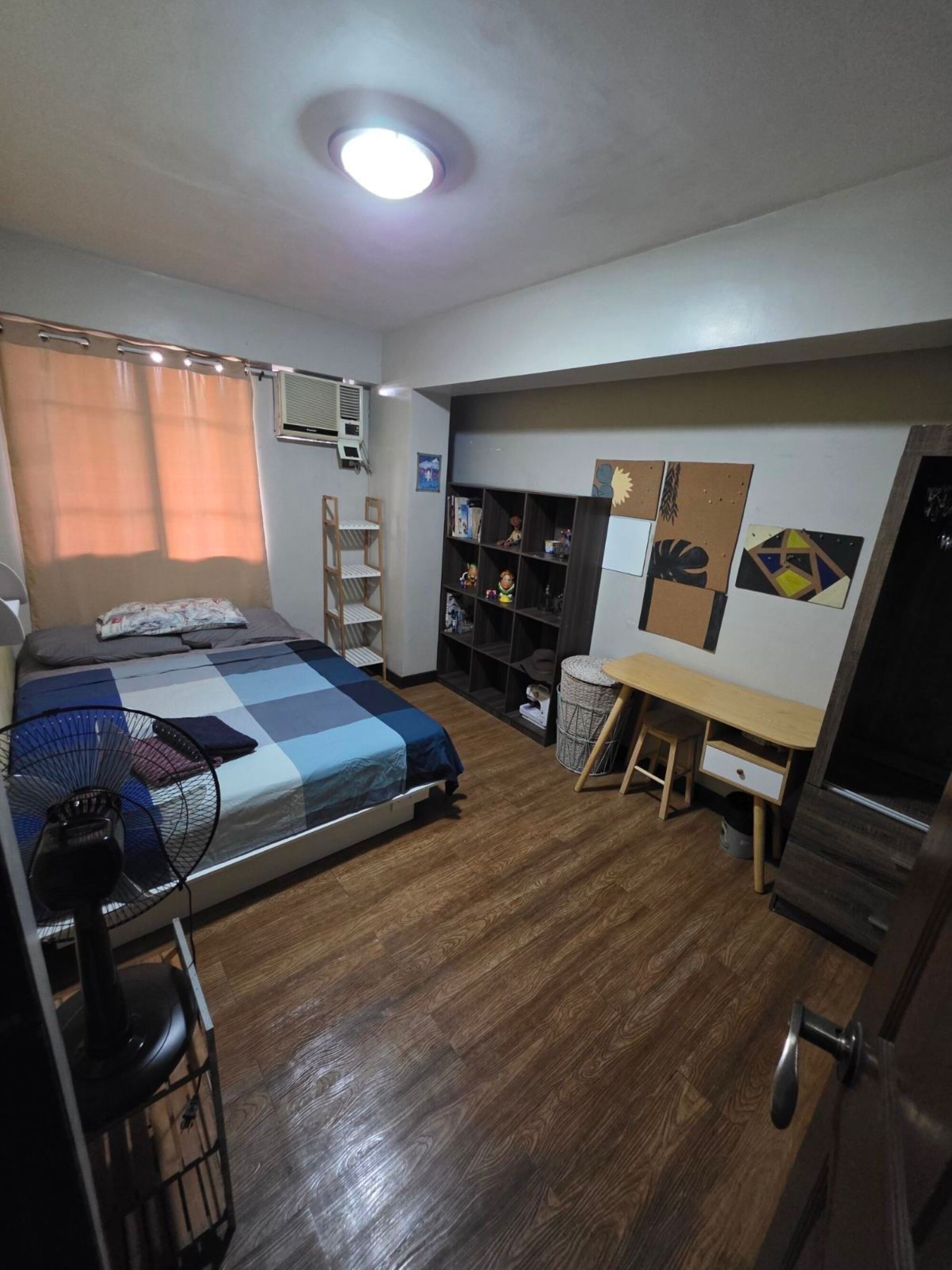 A compact bedroom with a bed, a desk, a chair, a fan, and a wardrobe.
