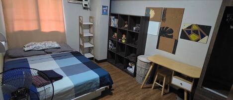 A compact bedroom with a bed, a desk, a chair, a fan, and a wardrobe.