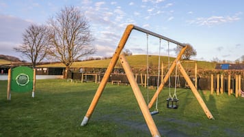 Un conjunto de columpios de madera en un césped, con un parque infantil cercado, un árbol y un edificio al fondo.