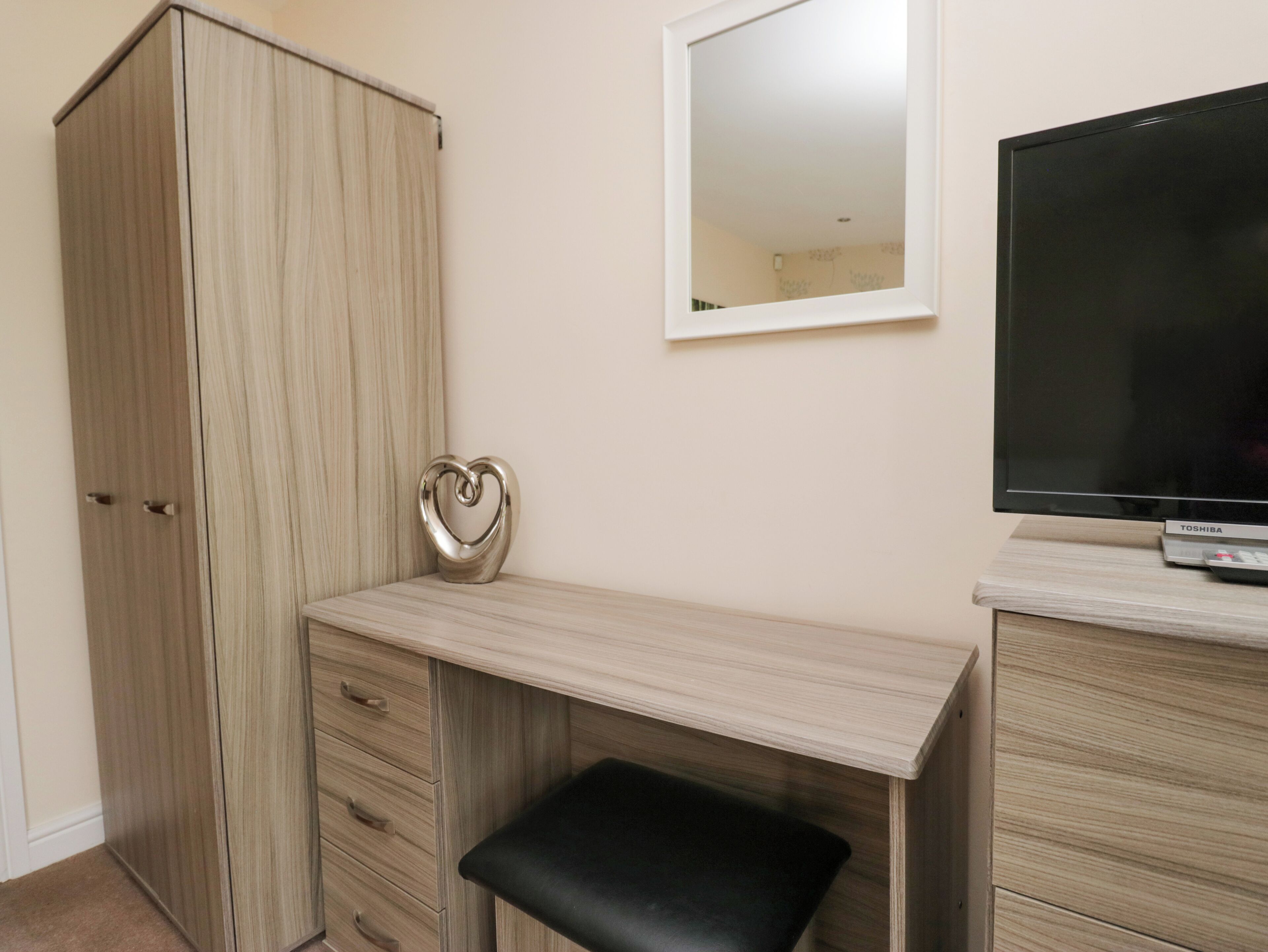 Een kamer met een kledingkast, een bureau met lades, een zwarte bank, een televisie en een spiegel aan de muur.