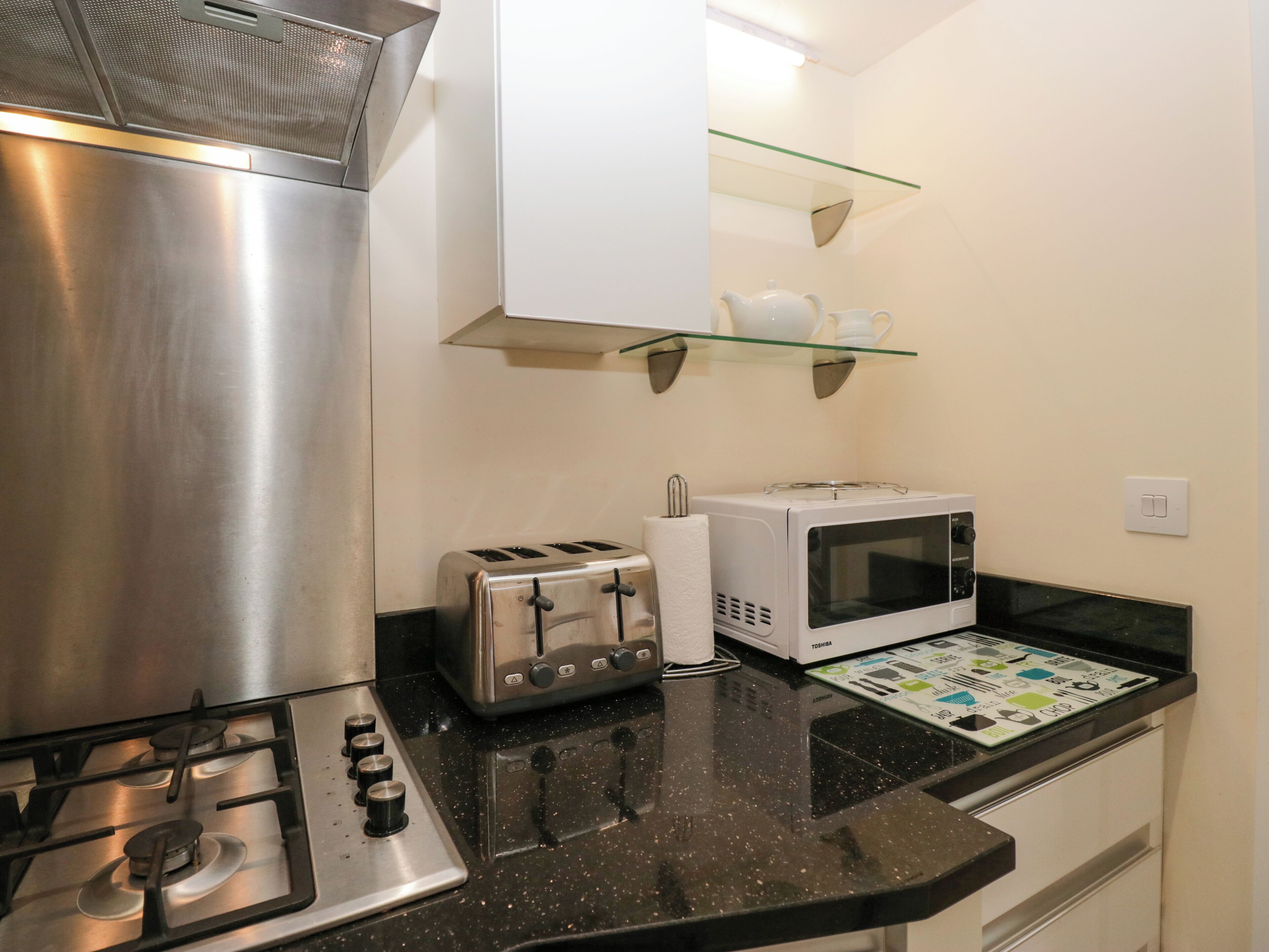 A compact kitchen with a gas stove, a toaster, a microwave, and a glass shelf above the counter.