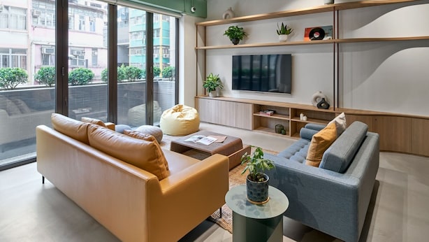 Un salon moderne avec un canapé, un fauteuil et une table basse, un téléviseur fixé au mur et de grandes fenêtres donnant sur des immeubles.