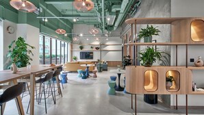 Un espace de bureau moderne avec une longue table, des chaises et une variété de tabourets colorés. On y trouve plusieurs plantes et une étagère en bois sur laquelle sont disposées d’autres plantes.