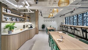 Un espace de bureau moderne avec un comptoir en bois aux lignes courbes, des chaises vertes et une longue table en bois. On y trouve des plantes en pot, une machine à café et une grande fenêtre donnant sur la ville.