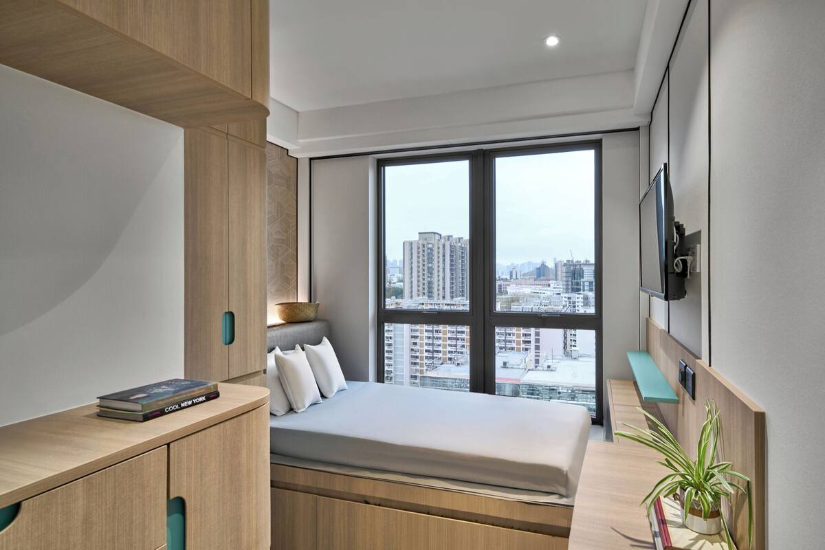 Une chambre moderne avec une grande fenêtre, un lit, un téléviseur fixé au mur et un bureau en bois agrémenté d’une plante.