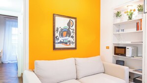 A living room with a white sofa, wooden flooring, a yellow wall, a ceiling with exposed beams, a hanging light fixture, and a bookshelf with decorative items.