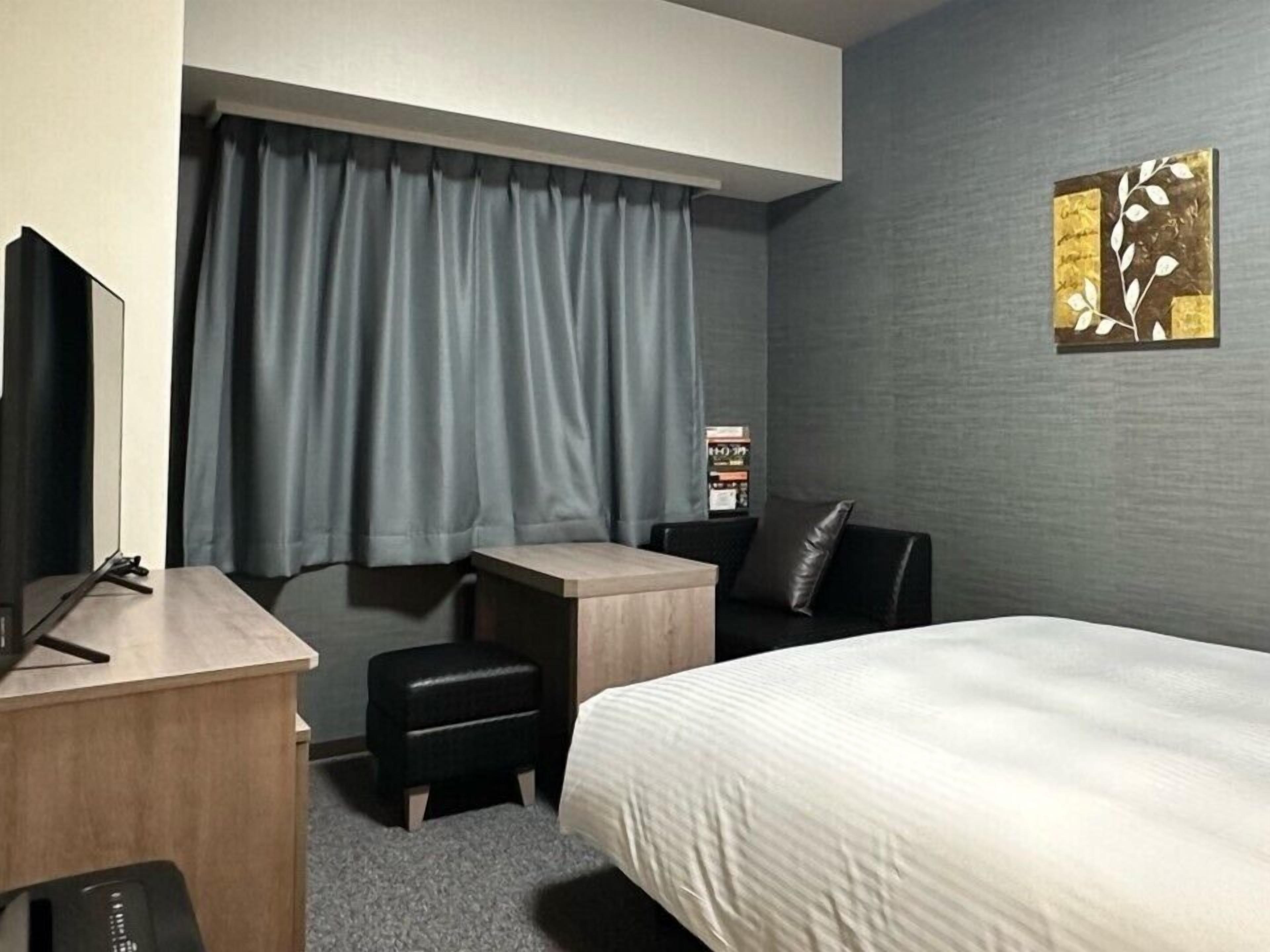 A hotel room with a bed, a desk, a chair, a small table, and a TV.