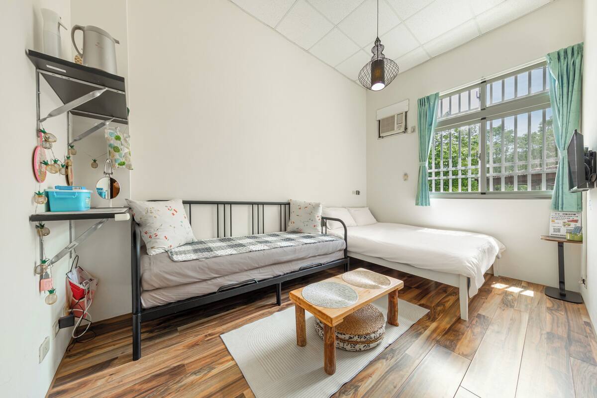 A compact room with a bed, a small table, a shelf with kitchen items, and a window with curtains.