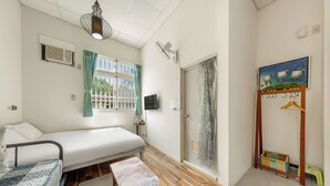 A compact room with a bed, a small table, a window with curtains, a wall-mounted fan, a shower area, and a painting. - Taomi Mountain Lodge (Puli)