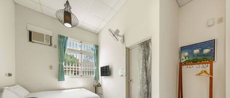 Habitación pequeña con cama, una mesita, ventana con cortinas, ventilador de pared, zona de ducha y un cuadro.