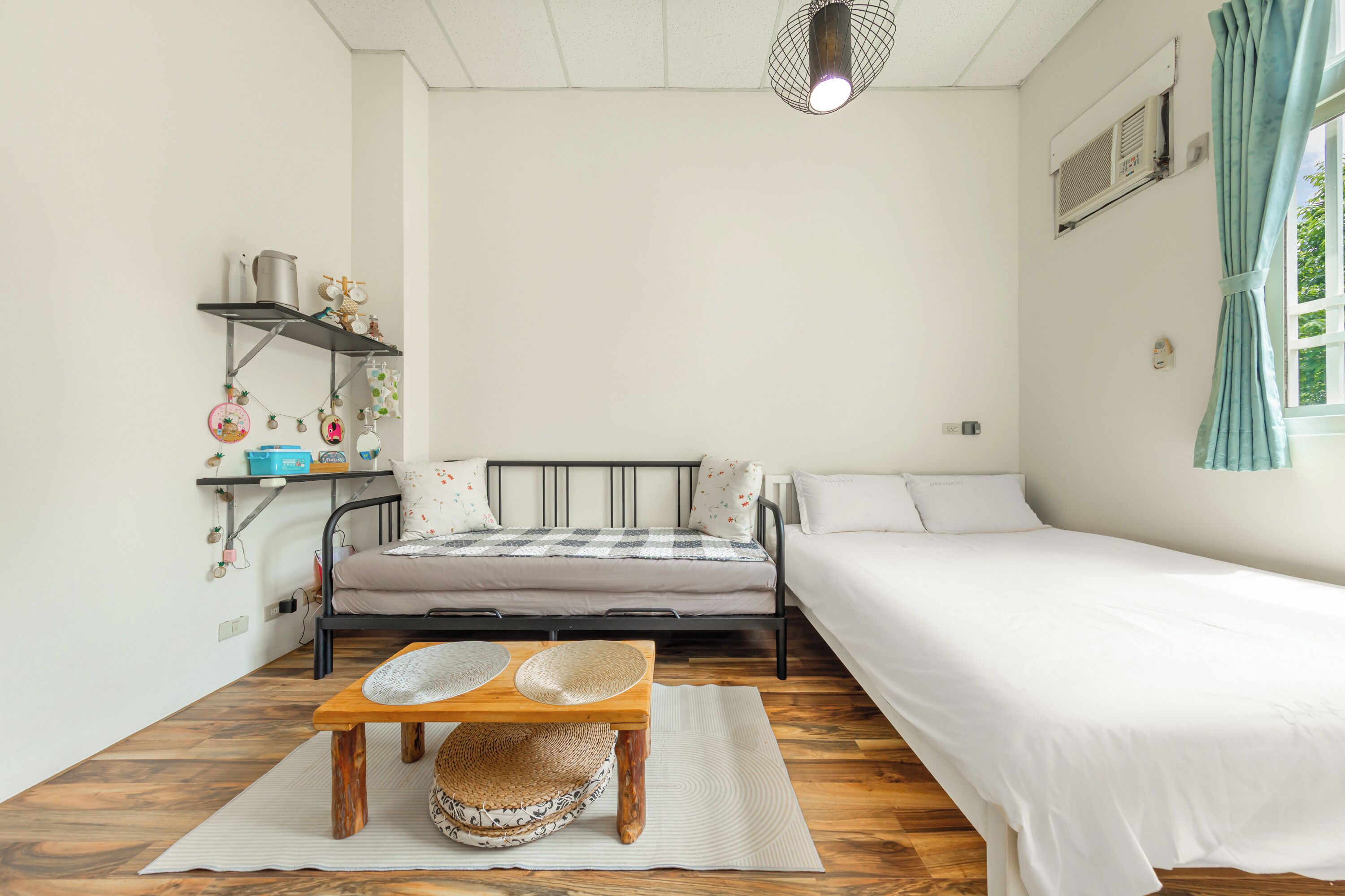 Quarto com cama, uma mesinha com cesta, prateleira com utensílios de cozinha e janela com cortinas.