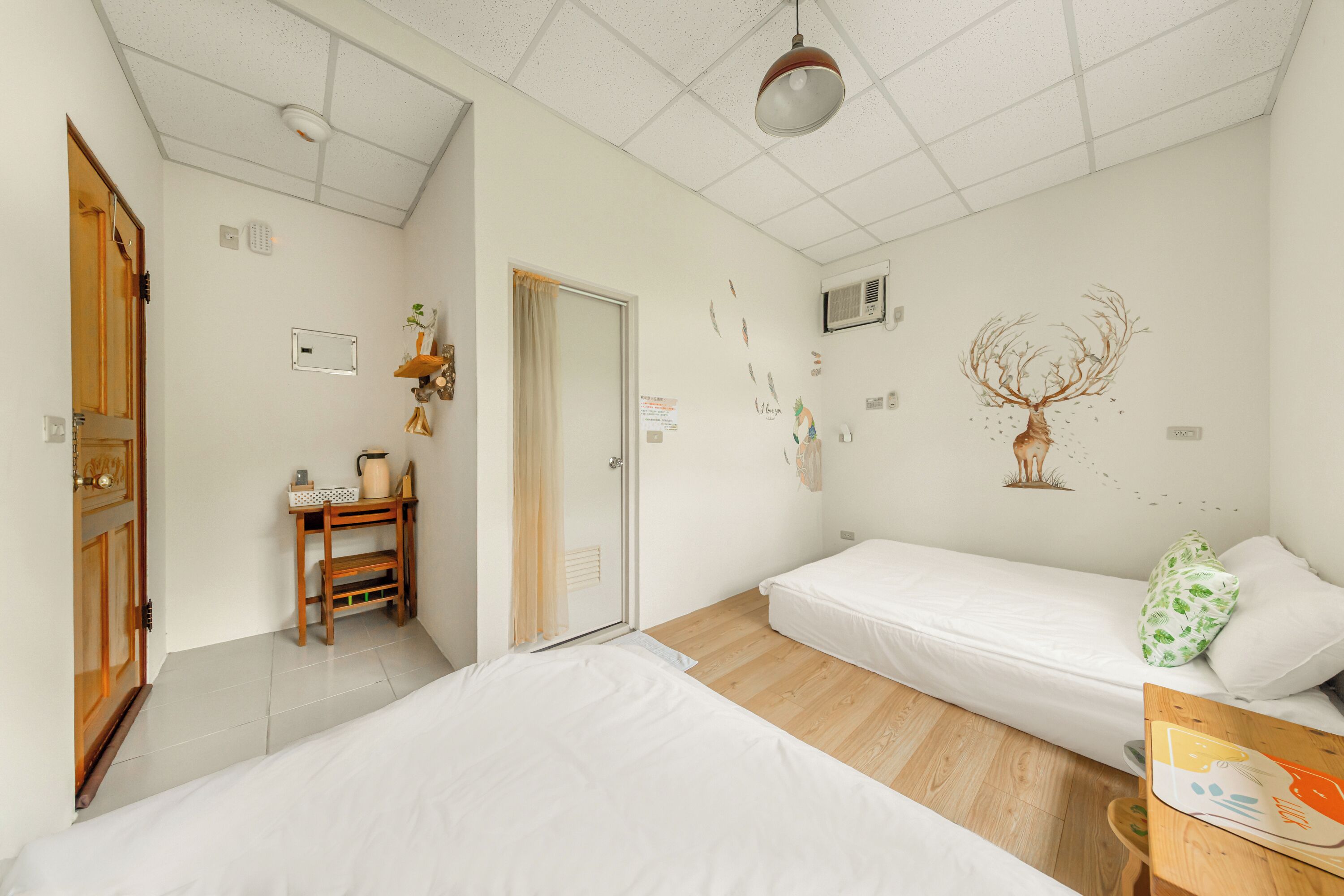 Un dormitorio con dos camas, un escritorio de madera, una silla, una mesita pequeña, un aire acondicionado de pared y una lámpara en el techo.