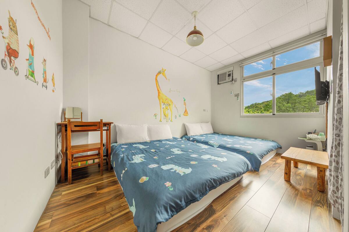 A bedroom with two beds, a wooden bench, a desk, and a window with a view of greenery.
