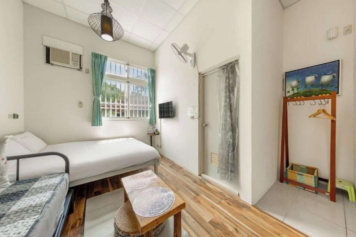 A small, well-lit room with a bed, a table, a window with curtains, a fan, and a painting on the wall.