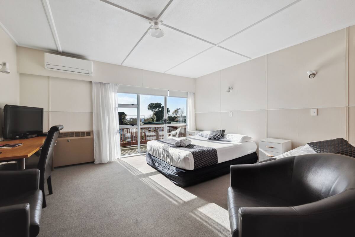 A modern hotel room with a large bed, a desk with a computer, and a balcony with a view.