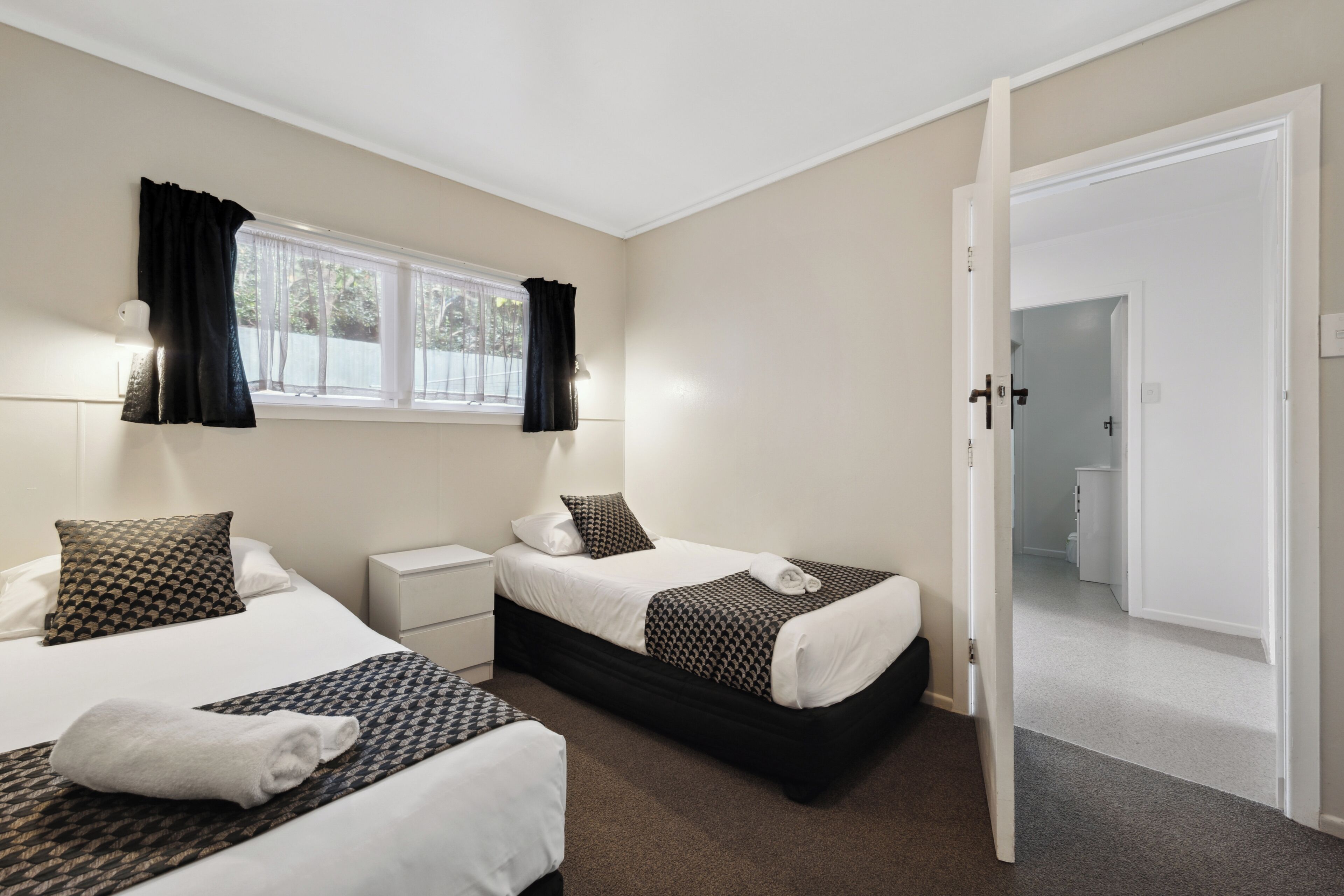 Una habitación de hotel con dos camas, una ventana con cortinas, una mesita de noche y una puerta que da a otra habitación.