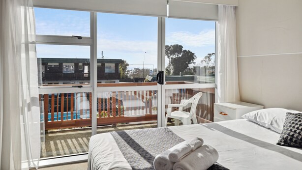 A hotel room with a large window, a bed with white linens, a small bedside table, and a view of a pool and buildings outside. - Flamingo Motel (New Plymouth)