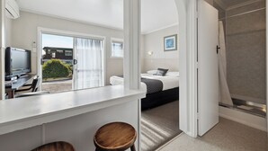 A modern hotel room with a kitchenette, a bed with a grey and black patterned bedspread, a shower area, and a view of the outside. - Flamingo Motel (New Plymouth)