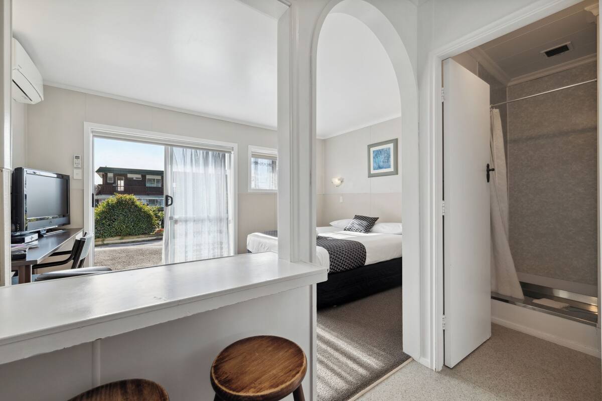 A modern hotel room with a kitchenette, a bed with a grey and black patterned bedspread, a shower area, and a view of the outside.