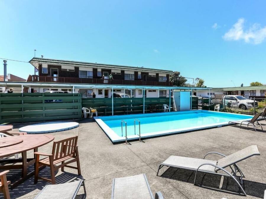 A swimming pool with lounge chairs and a table, surrounded by a fenced area with buildings in the background.